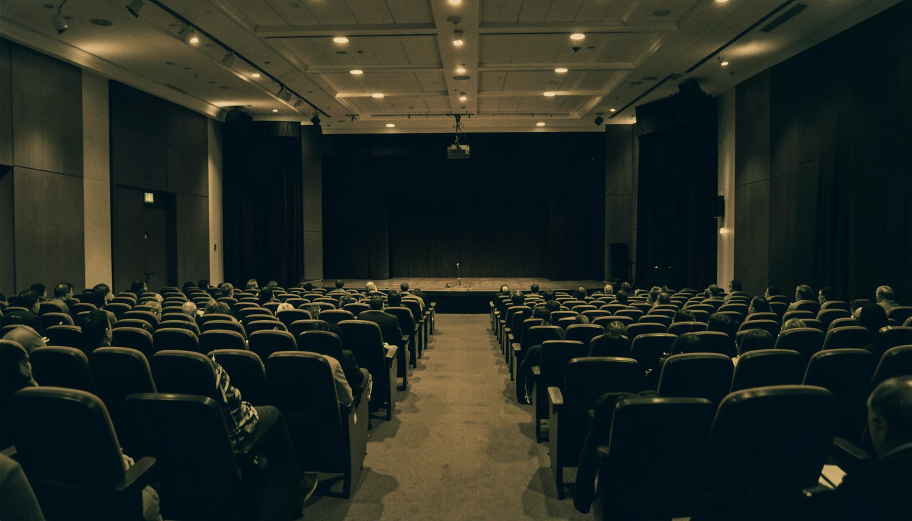 Duotone photograph of a conference hall interior with warm amber and deep navy tones, rows of seating and a stage