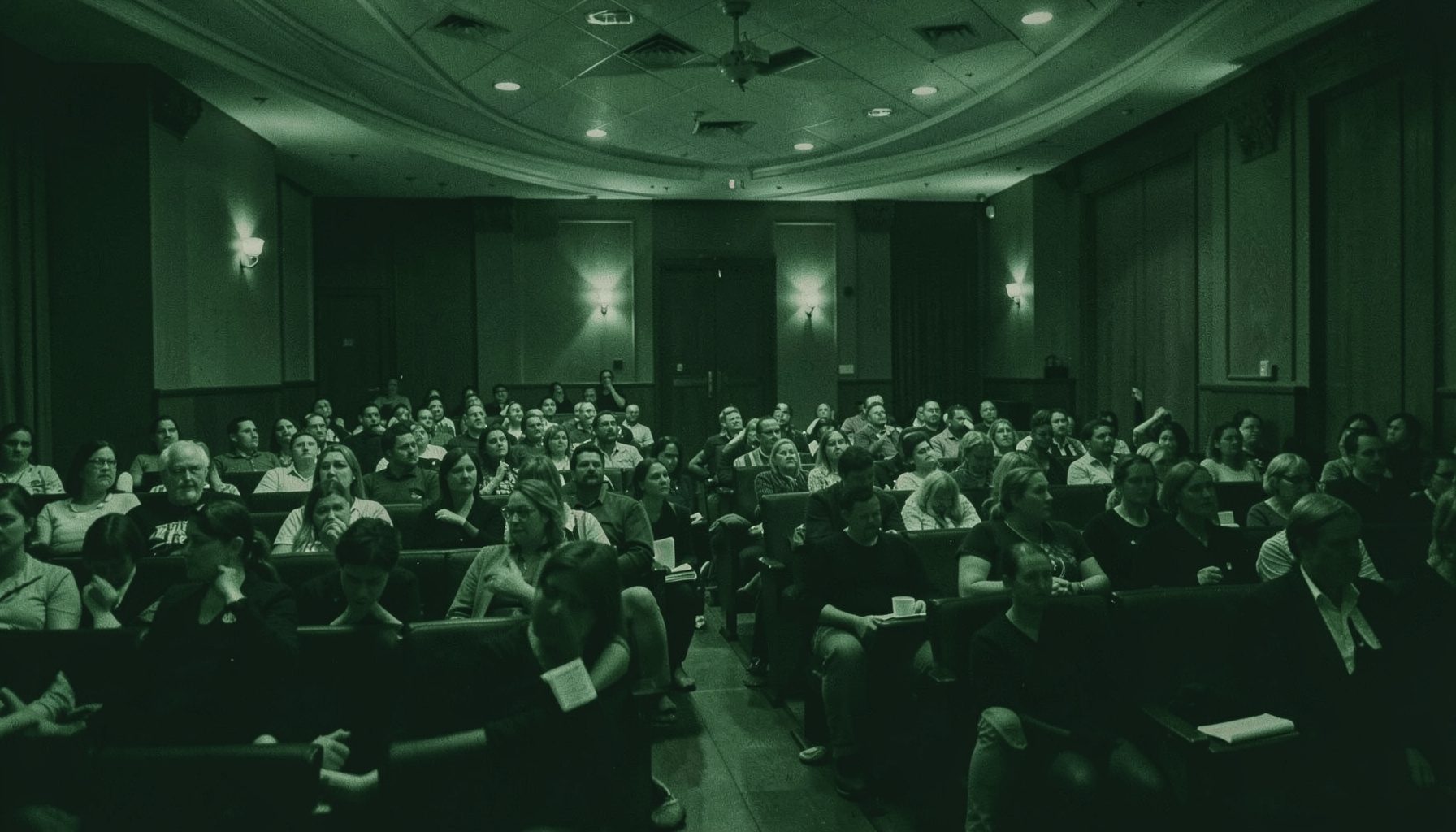 Deep green and charcoal duotone photograph of a lecture hall interior with warm lighting, suggesting an engaged academic audience